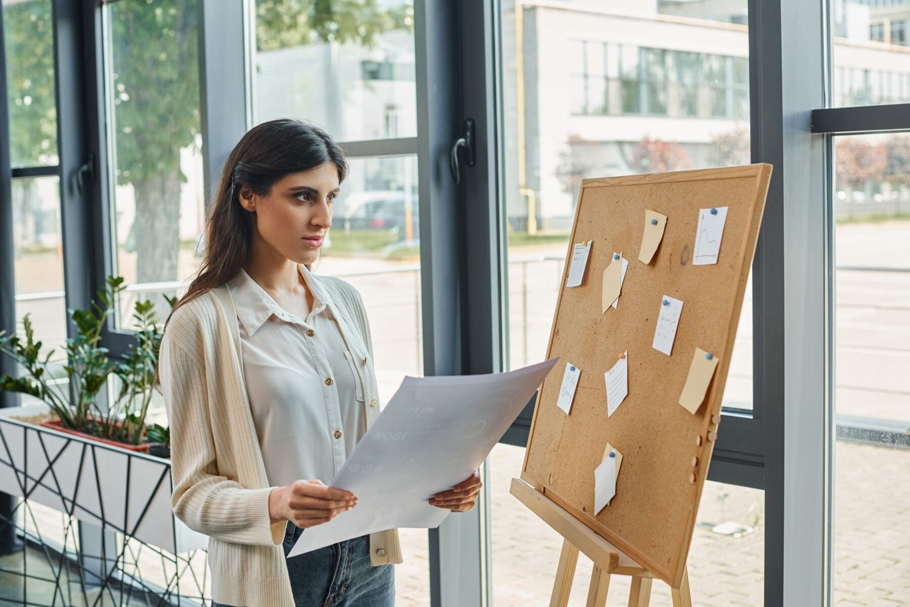 Calming Transformations Counseling - A lady wearing a silk white blouse and cardigan holding a A3 paper looking at pinboard for life coaching